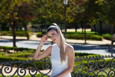 Uzun sarı saçlı ve kısa beyaz elbiseli güzel genç bir kadın Sevilla 'daki ünlü bir parkta, bitki örtüsü ve kuşlarla dolu bir bankta oturuyor. Seyahat ve tatil konsepti