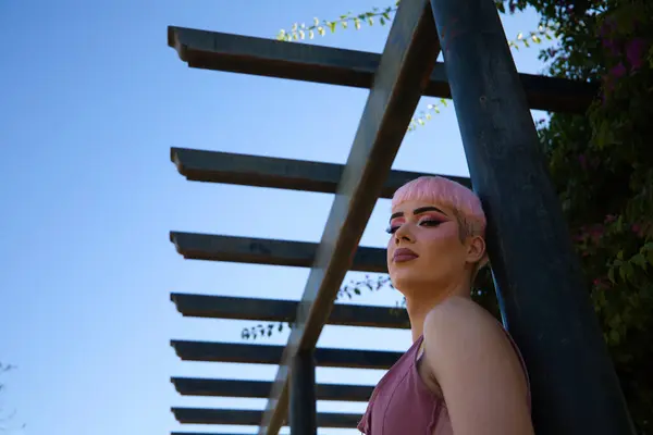 stock image Young gay guy with pink hair and make-up and dressed in pink is leaning on a poster in the park. The boy is sad and depressed because of the discrimination. Concept of equality and LGBTQ rights