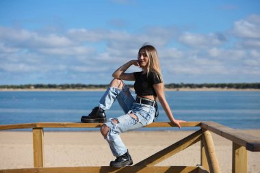 Yırtık kotlu, siyah bluzlu ve çizmeli güzel bir kız. Kız asi ve sert bir karakteri var. Kız plaj korkuluklarında oturuyor.