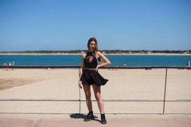 Siyah etek ve siyah bluz giymiş sarışın, genç ve güzel bir kadın denizin kenarında. Kız farklı vücut duruşları yapıyor. Arka planda mavi deniz