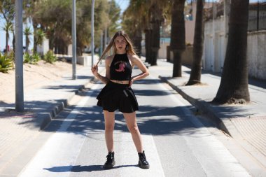 Siyah etek ve siyah bluz giymiş sarışın, genç ve güzel bir kadın yaya geçidinden geçiyor. Kadın karşıdan karşıya geçiyor ve farklı pozlar verme fırsatını yakalıyor.