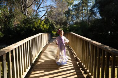 Fırfırlı ve püsküllü tipik bir çingene elbisesi içinde flamenko dansı yapan güzel bir kız İspanya 'nın Seville kentindeki ünlü bir parkta ahşap bir köprüde yürüyor. Saçları bir çiçekle bağlanmış.