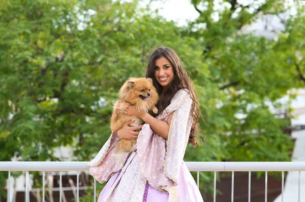 Beautiful Latin woman with long curly hair wearing a 15th century dress is holding a pomenarian dog in her arms. The woman is happy with her pet. Dog and pet day concept.