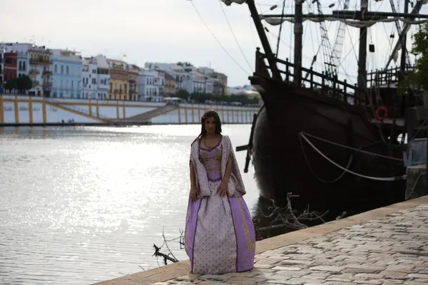 Beautiful Latin woman with long curly hair dressed in a 15th century dress next to one of the ships that discovered America in 1492. The ship is in the dock of seville in spain