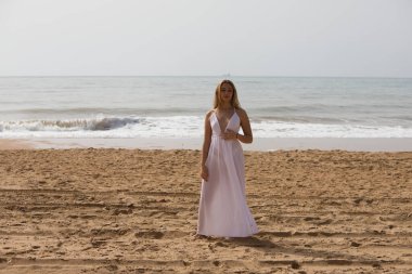 Beyaz elbiseli güzel sarışın kadın güneşli bir günde kumsalın kıyısında yürüyor. Kadın farklı vücut ifadeleri kullanıyor. Arka planda mavi deniz.