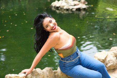 Genç ve güzel esmer kadın ve Latin, ördeğin taştan kenarında ve parktaki balık havuzunda oturuyor. Kadın gülümser, mutludur ve farklı ifadeler kullanır.