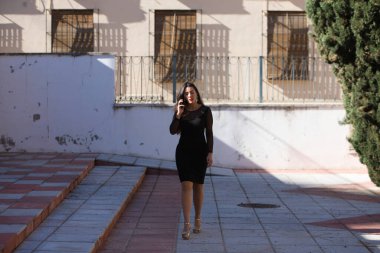 Genç, güzel esmer kadın siyah bir elbise giyiyor şeffaflığı ve süslü küpeleri var. Kız telefonda yürürken ve konuşurken yüz ifadeleri takınıyor.