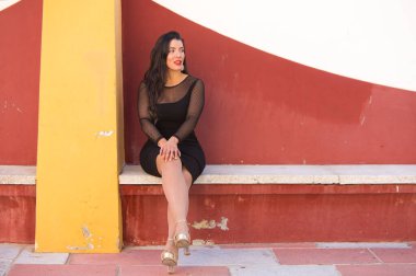 Genç, güzel esmer kadın siyah bir elbise giyiyor şeffaflığı ve süslü küpeleri var. Kız bir bankta bacak bacak üstüne atmış oturuyor. Güzellik ve moda konsepti