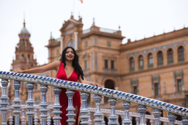 Kısa kırmızı elbiseler ve kırmızı ayakkabılar giymiş genç ve güzel esmer ve Latin bir kadın İspanya 'nın Seville Meydanı' nı ziyaret ediyor. Bir kadın fayanslı bir köprünün parmaklıklarına yaslanıyor ve mutlu..