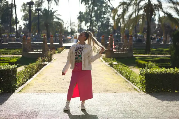 Beautiful young blonde woman with a ponytail in her hair wearing a pink skirt and fringed jacket walks through a famous park in Seville. The woman throws her head back and touches her hair
