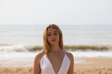 Beyaz elbiseli güzel sarışın kadın güneşli bir günde kumsalın kıyısında yürüyor. Kadın farklı vücut ifadeleri kullanıyor. Arka planda mavi deniz.