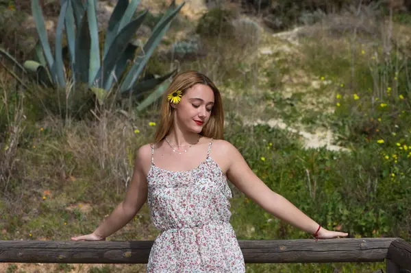Beautiful young blonde woman is wearing yellow daisy in her hair and is happy. The woman is wearing a dress with a flower pattern and is leaning on a wooden railing. Spring and nature concept.