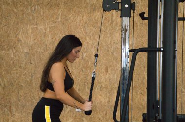 Genç ve güzel esmer kadın spor salonunda antrenman yapıyor. Kadın kol ve omuz egzersizleri yapıyor ve siyah bir bluz ve şort giyiyor. Sağlık ve spor kavramı