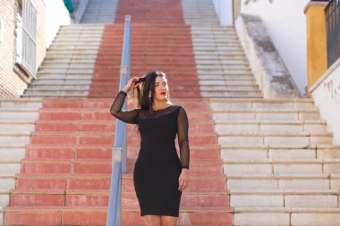 Genç, güzel esmer kadın şeffaflığı olan siyah bir elbise giyiyor. Kız caddede bir merdivenden aşağı yürüyor. Kadın kırmızı dudaklı makyaj yapıyor.