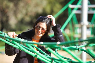 Genç, güzel, koyu renk saçlı, güneş gözlüklü ve siyah ceketli kadın çocuk parkının yeşil ipleri arasında. Kadın eğleniyor ve çok mutlu.