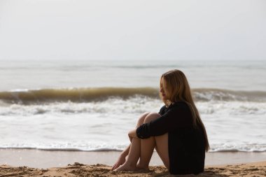 Siyah gömlekli güzel sarışın kadın sahilde üzgün ve depresif bir şekilde oturuyor. Kadın dalgın ve yere bakıyor bitkin ve yorgun. Kadın acı çekiyor..