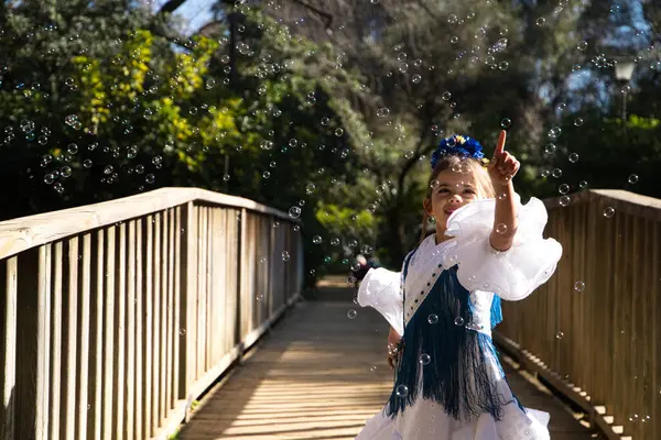 stock image A pretty girl dancing flamenco in a typical gypsy dress with frills and fringes walks on a wooden bridge in a famous park in Seville, Spain. The girl plays with soap bubbles.