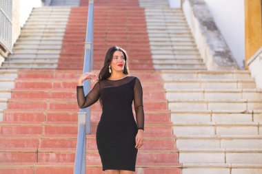 Genç, güzel esmer kadın şeffaflığı olan siyah bir elbise giyiyor. Kız caddede bir merdivenden aşağı yürüyor. Kadın kırmızı dudaklı makyaj yapıyor.