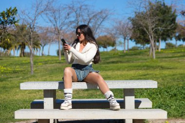 Parktaki bankta oturan genç ve güzel İspanyol esmer kadın. Kız rahat giyiniyor ve güneşli bahar gününün tadını çıkarırken cep telefonuna danışıyor.