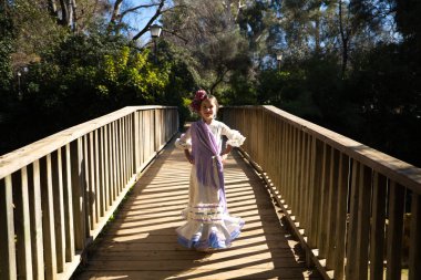 Fırfırlı ve püsküllü tipik bir çingene elbisesi içinde flamenko dansı yapan güzel bir kız İspanya 'nın Seville kentindeki ünlü bir parkta ahşap bir köprüde yürüyor. Saçları bir çiçekle bağlanmış.