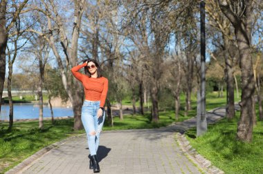 Gündelik kıyafetler ve güneş gözlüğü takmış genç ve güzel esmer kadın, sonbaharda yaprak döken ağaçlar arasında bir yol boyunca yürüyor, arka planda bir göl.