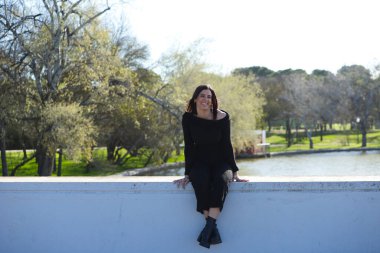 Genç ve güzel bir kadın, esmer ve siyah giyinmiş parkta bir duvarda oturuyor, arka planda sonbaharda ağaçlar ve şafakta güneş, kadın farklı vücut duruşları sergiliyor.