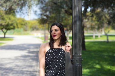 Güzel bir genç kadın, esmer ve siyah elbiseli, beyaz benekli, parkta bir sokak lambasına tutunmuş, kadın makyaj yapıyor ve kırmızı ruj sürüyor. Kadın mutlu ve gülümsüyor.