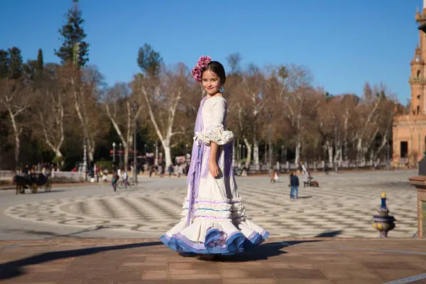 stock image a little girl dancing flamenco dressed in a beige dress with ruffles and purple fringes in a famous square in seville, spain. The girl has flowers on her head and her hair in a bun