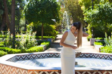 Uzun sarı saçlı ve kısa beyaz elbiseli güzel bir kadın Seville, İspanya 'da ünlü bir parkta. Arka planda bir çeşme, ağaçlar ve parkın heykelleri var. Seyahat ve tatil
