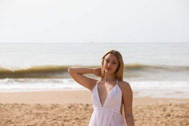 Beyaz elbiseli güzel sarışın kadın güneşli bir günde kumsalın kıyısında yürüyor. Kadın farklı vücut ifadeleri kullanıyor. Arka planda mavi deniz.