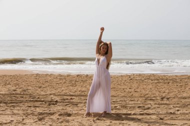 Beyaz elbiseli güzel sarışın kadın güneşli bir günde kumsalın kıyısında yürüyor. Kadın farklı vücut ifadeleri kullanıyor. Arka planda mavi deniz.