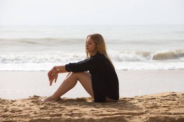 Siyah gömlekli güzel sarışın kadın sahilde üzgün ve depresif bir şekilde oturuyor. Kadın dalgın ve yere bakıyor bitkin ve yorgun. Kadın acı çekiyor..