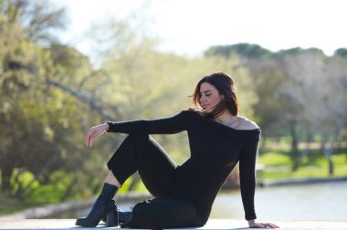 Genç ve güzel bir kadın, esmer ve siyah giyinmiş parkta bir duvarda oturuyor, arka planda sonbaharda ağaçlar ve şafakta güneş, kadın farklı vücut duruşları sergiliyor.