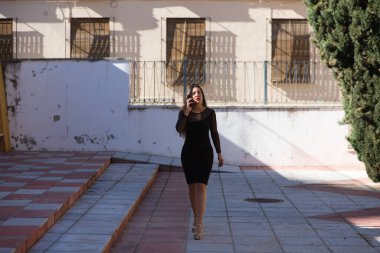 Genç, güzel esmer kadın siyah bir elbise giyiyor şeffaflığı ve süslü küpeleri var. Kız telefonda yürürken ve konuşurken yüz ifadeleri takınıyor.