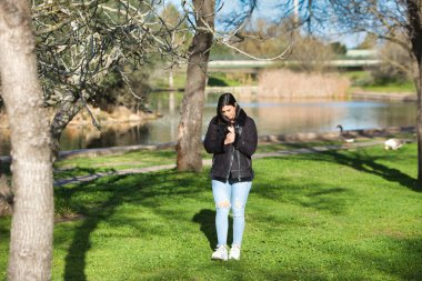 Kısa saçlı, kot pantolonlu ve siyah ceketli genç ve güzel esmer kadın soğuk bir günde yapraksız ağaçların arasında parkta yürürken kadın farklı vücut duruşları sergiliyor.
