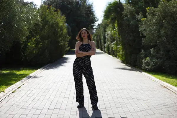 Genç, güzel, siyah elbiseli, beyaz puantiyeli esmer kadın kollarını çaprazlıyor ve önüne bakıyor, kadın makyaj yapıyor ve kırmızı ruj sürüyor.