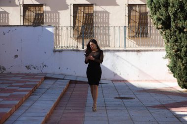 Genç, güzel esmer kadın siyah bir elbise giyiyor şeffaflığı ve süslü küpeleri var. Kız yürüyor ve telefonuna danışıyor. Kadın kırmızı dudaklı makyaj yapıyor.