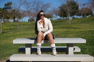 Parktaki bankta oturan genç ve güzel İspanyol esmer kadın. Kız rahat giyiniyor ve güneşli bahar gününün tadını çıkarırken farklı vücut ifadeleri takınıyor.