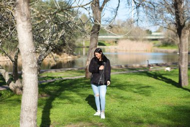 Kısa saçlı, kot pantolonlu ve siyah ceketli genç ve güzel esmer kadın soğuk bir günde yapraksız ağaçların arasında parkta yürürken kadın farklı vücut duruşları sergiliyor.