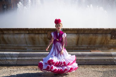 İspanya, Seville 'de ünlü bir meydanda pembe fırfırlar ve püsküllerle beyaz bir elbise içinde flamenko dansı yapan güzel bir kız. Kızın saçında bir çiçek var. Arka planda büyük bir çeşme var.