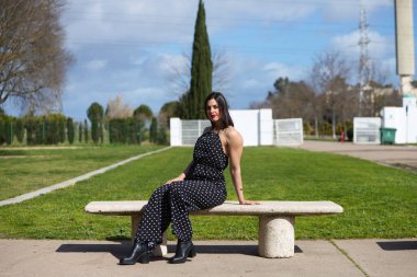 Güzel bir genç kadın, siyah elbiseli esmer, parktaki bankta beyaz benekli. Kadın mutlu ve güneşli günün tadını çıkarıyor.