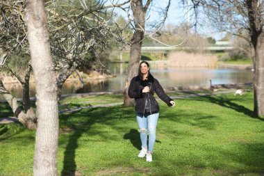 Kısa saçlı, kot pantolonlu ve siyah ceketli genç ve güzel esmer kadın soğuk bir günde yapraksız ağaçların arasında parkta yürürken kadın farklı vücut duruşları sergiliyor.