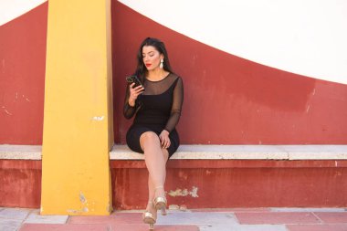 Genç, güzel esmer kadın siyah bir elbise giyiyor şeffaflığı ve süslü küpeleri var. Kız oturuyor ve ciddi bir yüzle telefonuna bakıyor.