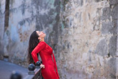 Makyajlı ve zarif kırmızı bir elbise giymiş güzel, genç İspanyol esmer kadın. Kız ellerini korkuluklara dayar ve saçı havada başını geriye doğru uzatır.