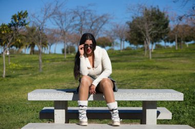 Parktaki bankta oturan genç ve güzel İspanyol esmer kadın. Kız rahat giyiniyor ve güneşli bahar gününün tadını çıkarırken farklı vücut ifadeleri takınıyor.