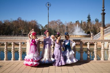 Seville, İspanya 'da ünlü bir meydanda tipik çingene kostümü giymiş dört güzel kız flamenko yapıyor. Flamenko, insanlığın kültürel mirası. Arka planda bir çeşme ve park var.