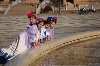Dört küçük kız tipik flamenko elbiseler içinde Flamenko dansı yapıyor Seville, İspanya 'daki ünlü bir meydanda çeşmeye yaslanıyorlar. Arkaplanda ünlü karenin kemerleri ve kolonları var.