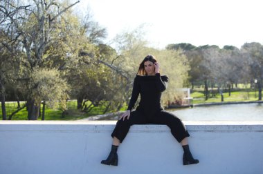 Genç ve güzel bir kadın, esmer ve siyah giyinmiş parkta bir duvarda oturuyor, arka planda sonbaharda ağaçlar ve şafakta güneş, kadın farklı vücut duruşları sergiliyor.