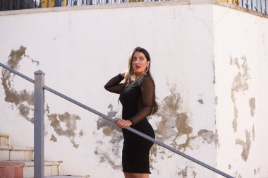 Genç, güzel esmer kadın şeffaflığı olan siyah bir elbise giyiyor. Kız sokakta bir merdivene tırmanıyor. Kadın kırmızı dudaklı makyaj yapıyor.