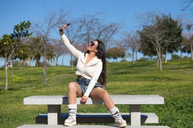 Parktaki bankta oturan genç ve güzel İspanyol esmer kadın. Kız rahat giyiniyor ve güneşli bahar gününün tadını çıkarırken cep telefonuyla selfie çekiyor.